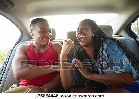 Stock Photograph - Young couple in car laughing, woman flexing muscles. Fotosearch