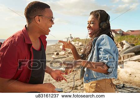 Stock Photo - Young couple on beach using headphones, standing face to face laughing. Fotosearch