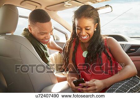 Stock Image - Young couple sitting in car looking at smartphone. Fotosearch