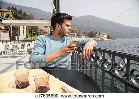 Stock Image - Young man looking out from lakeside restaurant, Lake Mergozzo, Verbania, Piemonte, Italy. Fotosearch