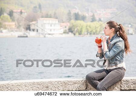 Stock Image - Young woman drinking cocktail on harbor wall, Lake Mergozzo, Verbania, Piemonte, Italy. Fotosearch