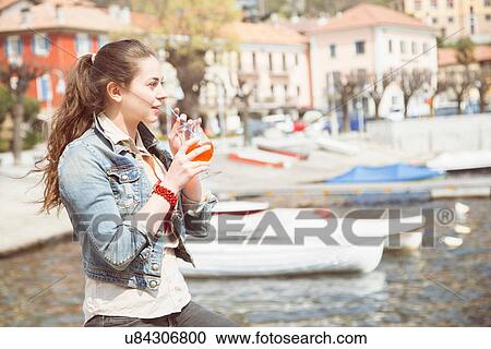 Stock Image - Young woman drinking cocktail on lakeside, Lake Mergozzo, Verbania, Piemonte, Italy. Fotosearch