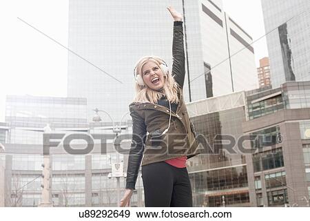 Stock Photo - Young woman on street jumping whilst listening to headphones, New York, USA. Fotosearch