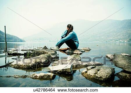 Picture - Young woman sitting on rock looking out over Lake Mergozzo, Verbania, Piemonte, Italy. Fotosearch