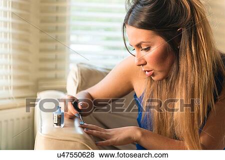 Stock Photo - Young woman sitting on sofa, painting nails. Fotosearch