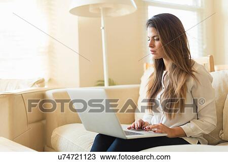 Stock Image - Young woman sitting on sofa, using laptop. Fotosearch