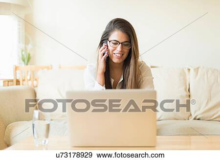 Young woman sitting on sofa, using laptop and talking on mobile phone View Large Photo Image Stock Photo - Young woman sitting on sofa, using laptop and talking on mobile phone. Fotosearch