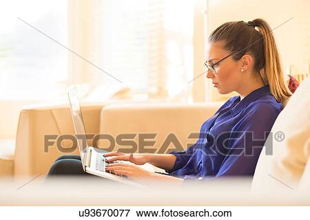 Stock Photo - Young woman sitting on sofa, using laptop. Fotosearch