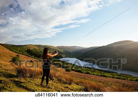Stock Image - Young woman taking smartphone photograph of Talybont Reservoir in Glyn Collwn valley, Brecon Beacons, Powys, Wales. Fotosearch