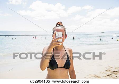 Stock Photography - Young woman wearing bikini top taking smartphone selfie on Waikiki Beach, Hawaii, USA. Fotosearch