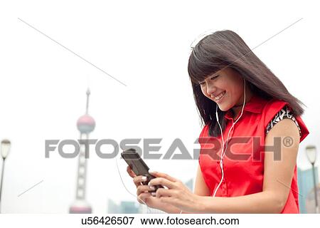 Stock Photo - Young woman wearing earphones, looking at smartphone, Shanghai, China. Fotosearch