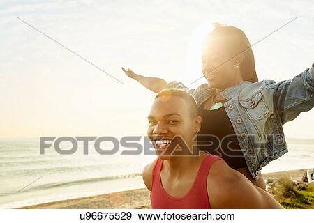Stock Photo - Young woman with arms raised standing behind young man, looking at camera. Fotosearch