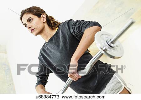 Stock Image - Angled view of young woman lifting barbell in living room. Fotosearch