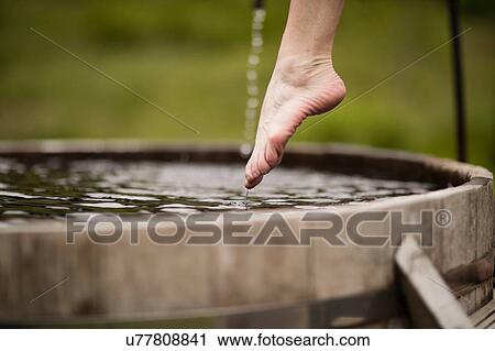 Stock Image - Bare foot of mature woman stepping into fresh cold water tub at eco retreat. Fotosearch