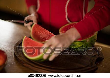 Stock Photograph - Cropped view of mature woman slicing watermelon in kitchen. Fotosearch