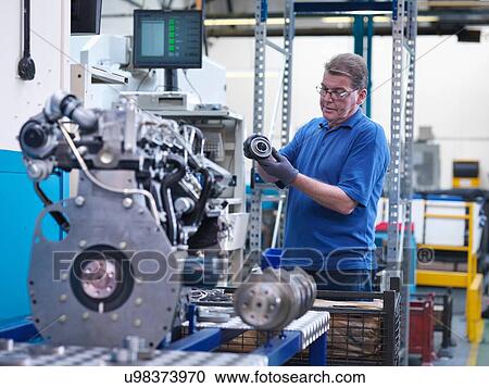 Stock Image - Engineer with gear wheel and diesel engine in engineering factory. Fotosearch