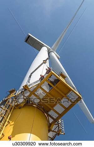 Stock Image - Engineers climbing wind turbine from boat at offshore windfarm, low angle view. Fotosearch