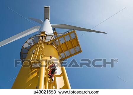 Stock Image - Engineers climbing wind turbine from boat at offshore windfarm, low angle view. Fotosearch