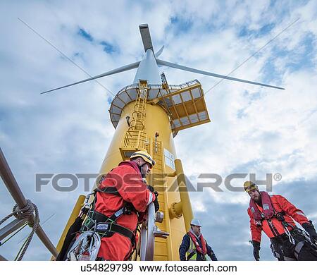 Stock Photo - Engineers climbing wind turbine from boat at offshore windfarm, low angle view. Fotosearch