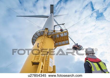Stock Photography - Engineers winch tools up wind turbine from boat at offshore windfarm, low angle view. Fotosearch