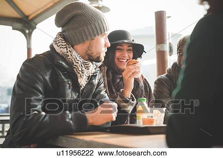Stock Image - Four young adult friends chatting and eating doughnuts at sidewalk cafe. Fotosearch