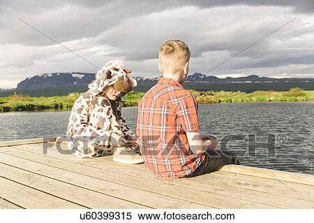 Stock Photography - Girl wearing animal onesie sitting on lake pier with brother, Thingvellir, Iceland. Fotosearch