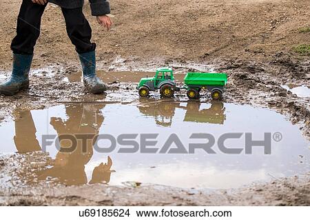 Picture - Legs of male toddler wearing rubber boots playing with toy tractor in muddy puddle. Fotosearch