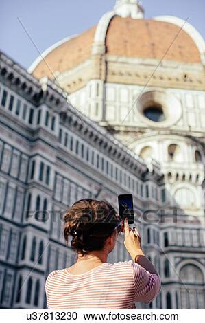 Stock Image - Low angle rear view of mid adult woman using smart phone to take photograph, Duomo Cathedral, Florence, Italy. Fotosearch