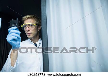 Stock Photo - Male scientist holding up and examining specimen in lab cleanroom. Fotosearch