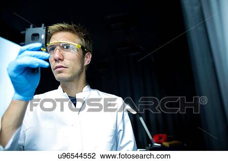 Stock Image - Male scientist holding up and scrutinizing specimen in lab cleanroom. Fotosearch