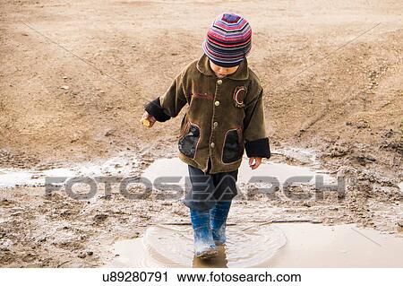 Stock Image - Male toddler wearing rubber boots paddling in puddle. Fotosearch