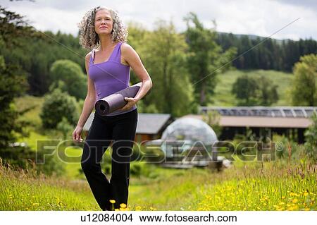 Mature woman carrying yoga mat walking in eco lodge field View Large Photo Image Picture - Mature woman carrying yoga mat walking in eco lodge field. Fotosearch