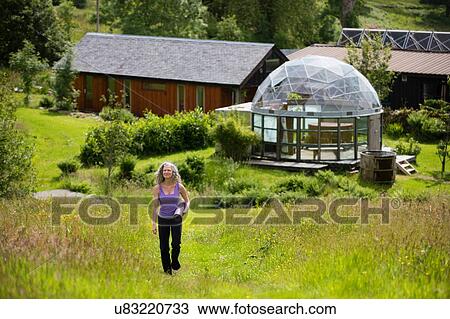 Stock Image - Mature woman carrying yoga mat walking up eco lodge field. Fotosearch