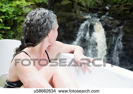 Picture - Mature woman in bubble bath gazing at waterfall at eco retreat. Fotosearch