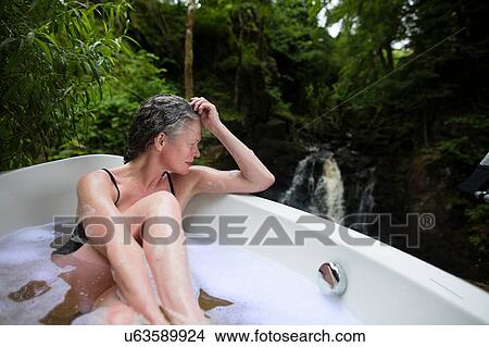 Picture - Mature woman in bubble bath in front of waterfall at eco retreat. Fotosearch