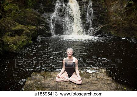 Stock Image - Mature woman practicing lotus pose in front of waterfall. Fotosearch