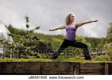 Stock Image - Mature woman practicing warrior pose in eco lodge garden. Fotosearch