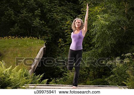 Stock Image - Mature woman practicing yoga pose on one leg in garden. Fotosearch