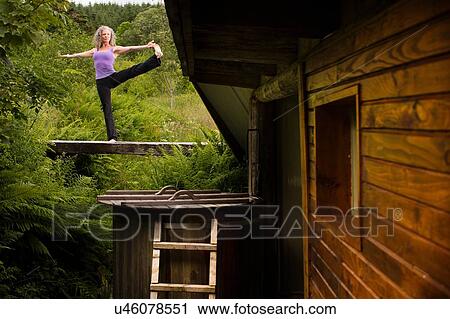 Stock Image - Mature woman practicing yoga pose on footbridge at eco lodge. Fotosearch