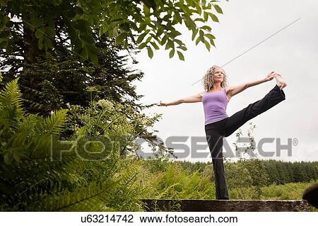 Stock Image - Mature woman practicing yoga pose on footbridge. Fotosearch