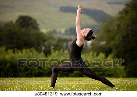 Stock Photo - Mature woman practicing yoga warrior pose with arms raised in field. Fotosearch