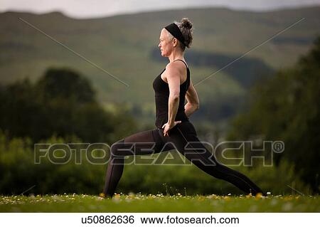 Stock Photograph - Mature woman practicing yoga warrior pose in field. Fotosearch