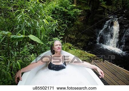 Stock Image - Mature woman relaxing in bubble bath in front of waterfall at eco retreat. Fotosearch