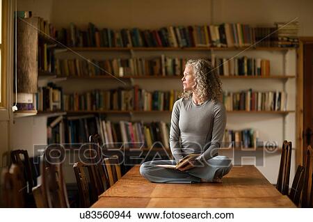 Picture - Mature woman sitting cross legged on kitchen table looking out of window. Fotosearch