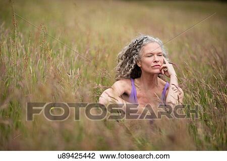 Stock Image - Mature woman sitting in long grass looking away. Fotosearch