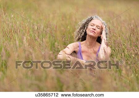 Stock Image - Mature woman sitting in long grass with eyes closed. Fotosearch