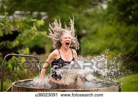 Stock Image - Mature woman splashing into fresh cold water tub at eco retreat. Fotosearch