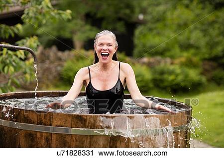 Stock Image - Mature woman standing in fresh cold water tub at eco retreat. Fotosearch
