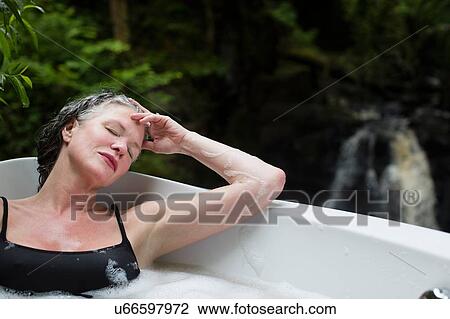 Stock Image - Mature woman with eyes closed in bubble bath in front of waterfall at eco retreat. Fotosearch