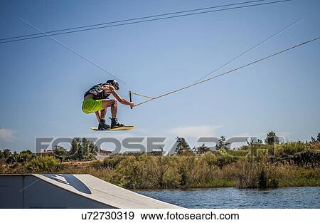 Stock Photo - Mid adult male wakeboarder jumping ramp in sea, Cagliari, Sardinia, Italy. Fotosearch
